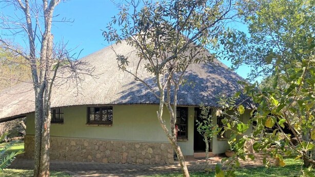 A building with a stone foundation and a thatched roof, surrounded by a grassy area and trees. - Family Lodge in Natural African Bush - 2115 (Bulawayo)