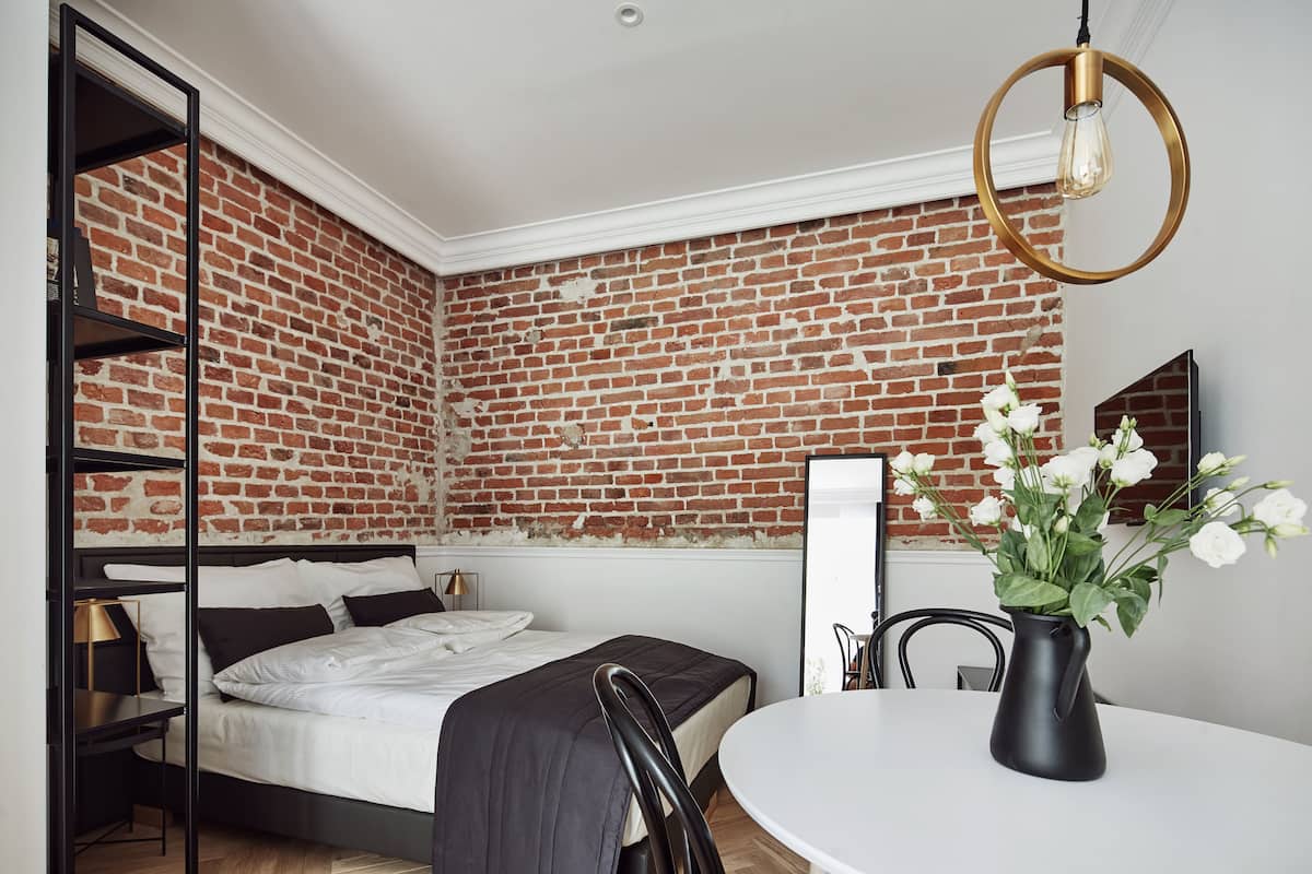 Um quarto com parede de tijolos, uma cama com cabeceira preta, uma mesa redonda com um vaso de flores e um lustre.