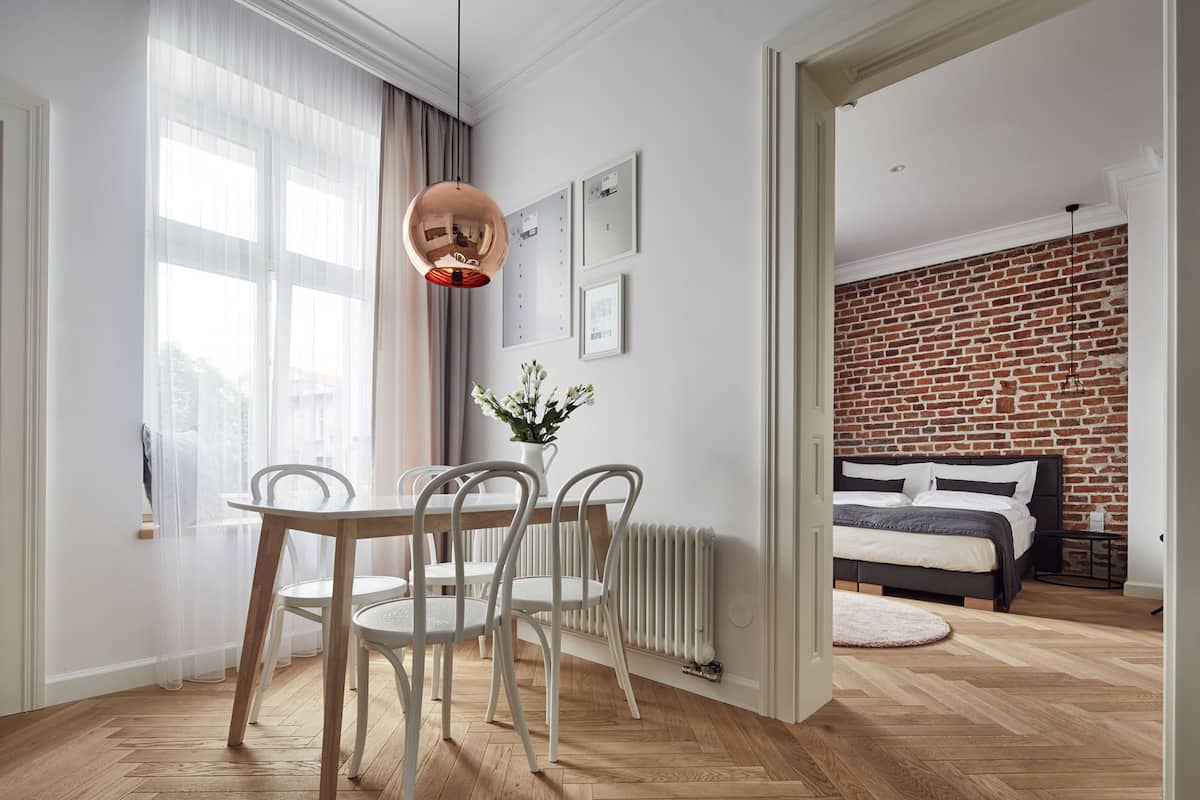 Quarto de hotel moderno com piso de madeira, área de refeições com cadeiras brancas, luminária pendente, parede de tijolos e cama com cabeceira preta.