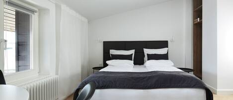 A modern bedroom with a black bed, white bedding, a small bedside table, and a window with blinds.
