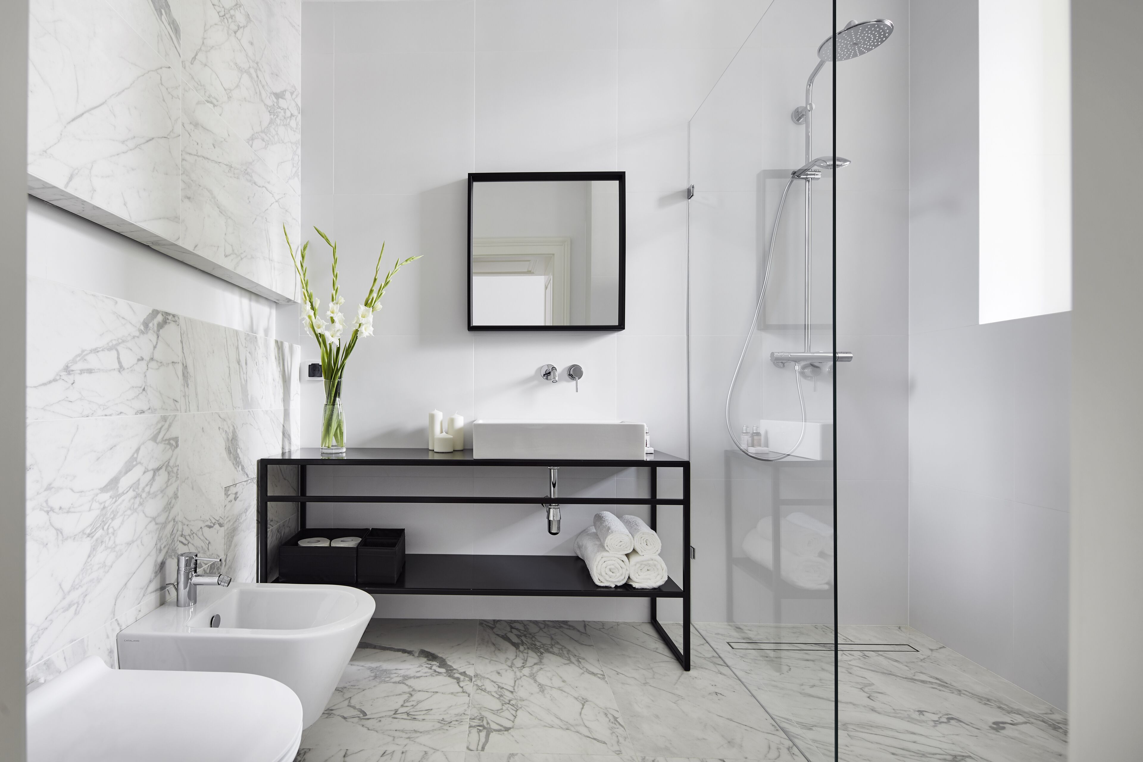 A modern bathroom with a marble floor, a freestanding bathtub, a black vanity with a mirror, a glass-enclosed shower, and a vase with flowers on the vanity.