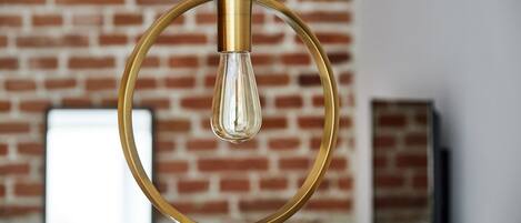 A hanging light fixture with a clear glass bulb encased in a gold-colored circular frame, set against a brick wall background.