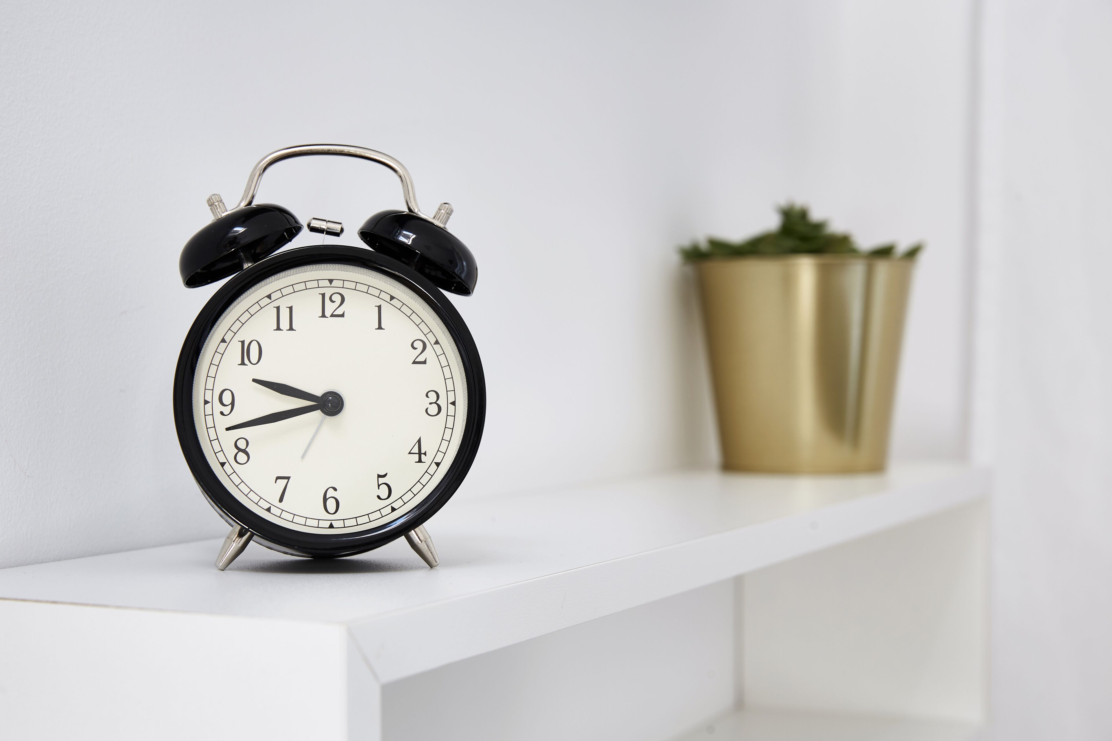 A black analog alarm clock on a white shelf with a gold pot in the background.