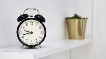 A black analog alarm clock on a white shelf with a gold pot in the background.