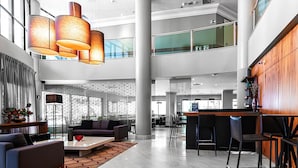 Een ruime lobby met een hoge plafond, moderne verlichtingsarmaturen, een zithoek met sofa’s, een salontafel en een bar met barkrukken.