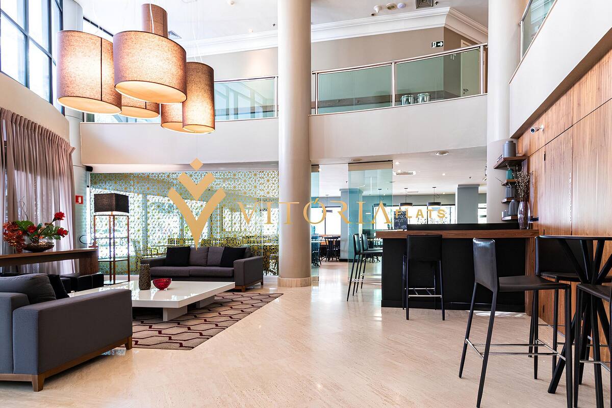 Um lobby amplo com design moderno, apresentando uma área de estar com sofás e uma mesa de centro, uma área de bar com banquetas, e uma grande letra decorativa 'Y' na parede.