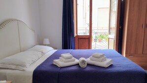 A neatly made bed with white linens and a blue blanket, a wooden nightstand with a lamp, and a view of a balcony with potted plants. - Appartamenti Blu Mare "SCOPELLO" situeted in a strategic position (Castellammare del Golfo)