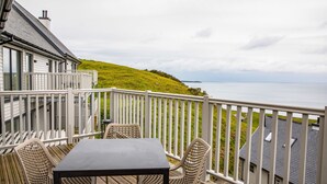 Eine Terrasse mit Tisch und Stühlen, mit Blick auf einen grasbewachsenen Hügel und das Meer.