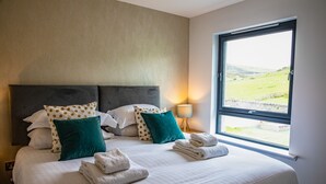 Ein ordentlich bezogenes Bett mit weißen Leinen und grünen Kissen, ein Fenster mit Blick auf eine grüne Landschaft und eine Nachttischlampe.