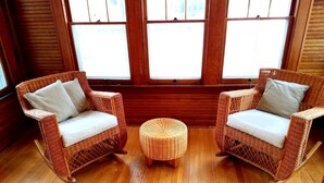 Two wicker chairs with cushions, a small round ottoman, and wooden flooring in a room with large windows. - Franklin Manor Bed & Breakfast (Saranac Lake)