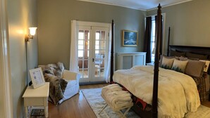 A bedroom with a four-poster bed, a white nightstand with a framed menu, a chair with a fur throw, and a view through a glass door to a balcony. - Franklin Manor Bed & Breakfast (Saranac Lake)