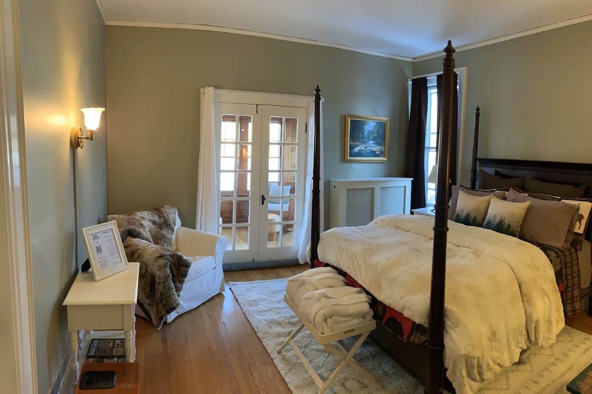 A bedroom with a four-poster bed, a white nightstand with a framed menu, a chair with a fur throw, and a view through a glass door to a balcony.
