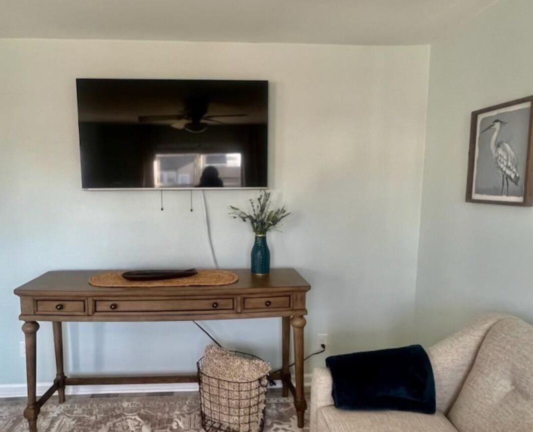 Un salon avec un téléviseur fixé au mur, une table en bois avec des tiroirs, un vase avec des plantes et un canapé avec un coussin.