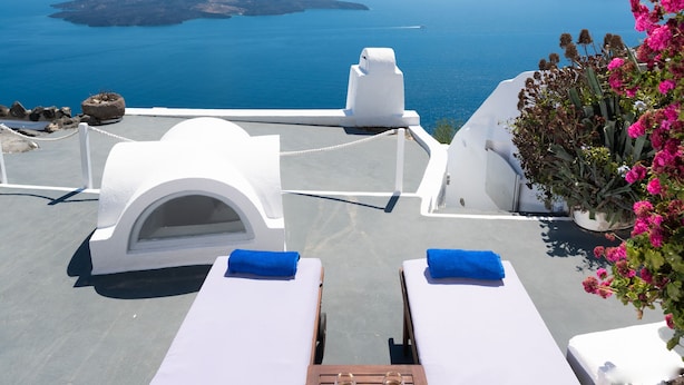 A sunlit outdoor seating area with two lounge chairs and a table, overlooking a scenic view of the sea and islands. - Iatis View Collection (Santorini)