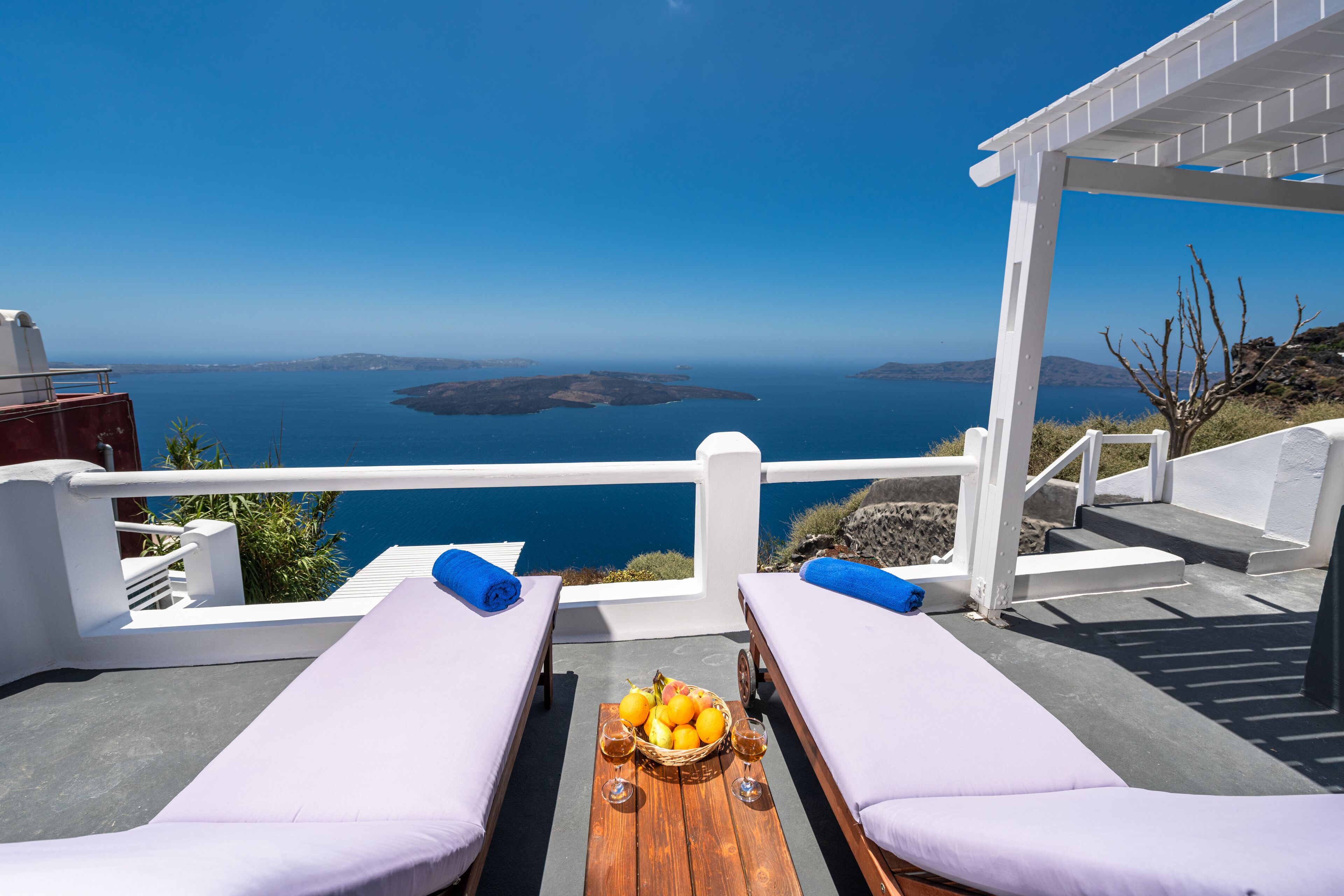A balcony with two sunbeds and a table with fruit and drinks, overlooking a scenic view of the sea and islands.