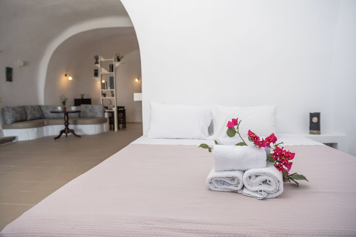 Ein ordentlich bezogenes Bett mit aufgerollten weißen Handtüchern und rosa Blumen, eine weiße Wand und Blick auf einen Wohnbereich mit Sofa und Tisch.