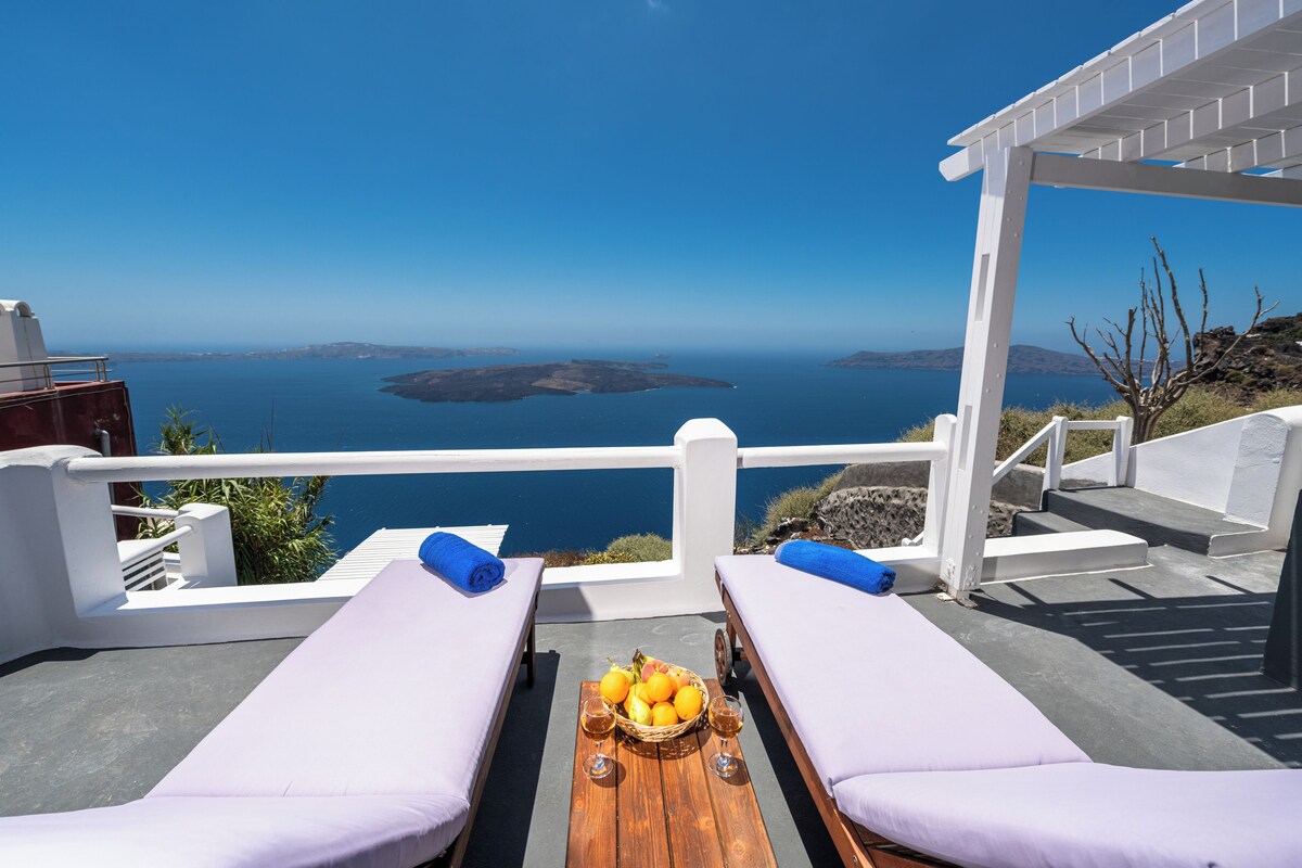 Eine Strandterrasse mit zwei Liegestühlen, einem Holztisch mit Obst und Weingläsern und Blick auf das Meer und Inseln.