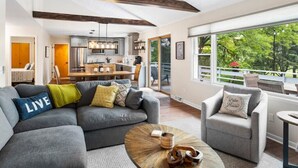 A modern living room with a gray sectional sofa, a round coffee table, and a gray armchair. A kitchen is visible in the background with stainless steel appliances.
