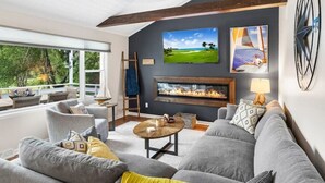 A modern living room with a fireplace, grey sofas, and a large window overlooking greenery.