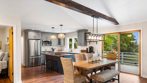 A modern kitchen with stainless steel appliances, a dining area with wicker chairs, and large windows offering a view of the outdoors.