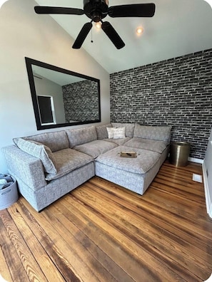 Un salon moderne avec un canapé d’angle, un ventilateur de plafond, un mur décoré d’un motif en briques et un plancher en bois.