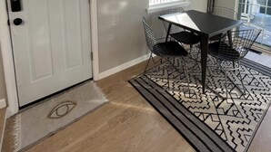 Een kamer met een witte deur, een raam met zonwering, een eettafel met stoelen en een kamerbreed tapijt.