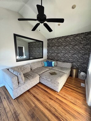 A modern living room with a sectional sofa, a ceiling fan, a brick-patterned wall, and a wooden floor.