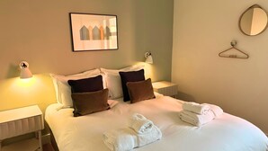 Een net opgemaakt bed met kussens, een bedlampje, een ingelijst plaatje, een spiegel en een stap handdoeken.