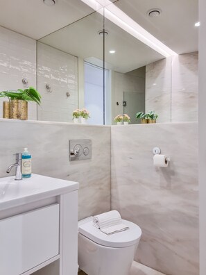 A modern bathroom with a white sink, a toilet, and a large mirror. There are plants on the sink and a towel on the toilet.