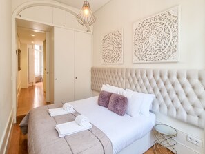 A bedroom with a tufted headboard, white bedding, a decorative wall piece, and a bedside table with a lamp.