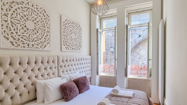 Una cama bien tendida con un cabecero capitonado, almohadas blancas y rosas, y una colcha color beige. Hay paneles decorativos en la pared sobre la cama y una vista al exterior a través de dos ventanas.