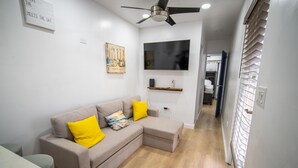 A living room with a beige sectional sofa, two yellow pillows, a flat-screen TV, a ceiling fan, and a wooden floor. - Sunset Villas "Good Morning" Tiny Home Container (Frederiksted)