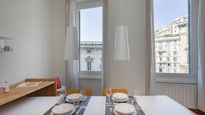 A dining area with a table set for two, two chairs, a wooden sideboard with a bottle and a menu, and a view of a building through a window. - Cairoli Stylish Apartment by Wonderful Italy (Genoa)