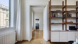 A room with a wooden floor, white walls, and a large window. There is a bookshelf with books and a small table with a bowl and a bottle. - Cairoli Stylish Apartment by Wonderful Italy (Genoa)