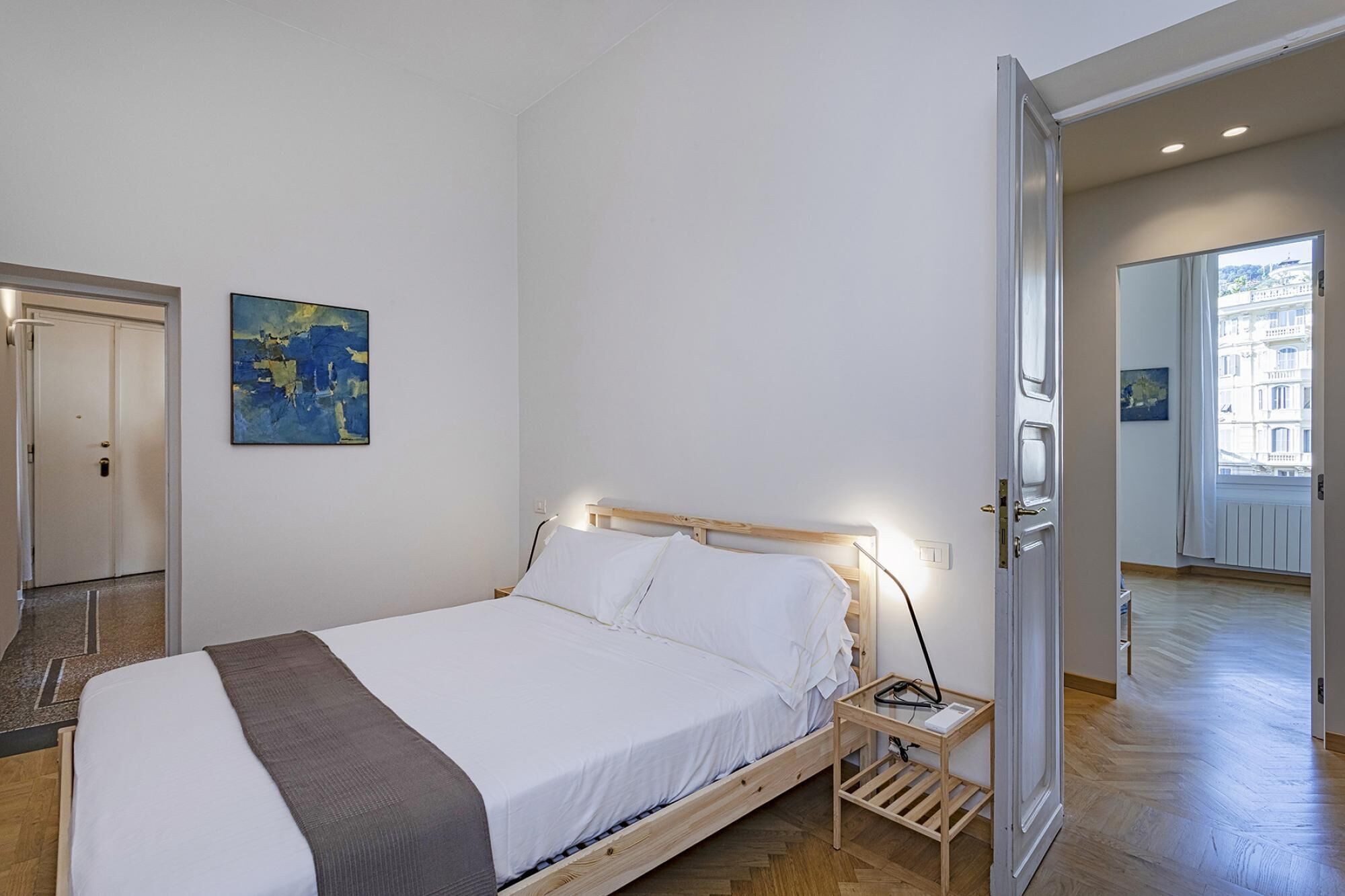 Ein Schlafzimmer mit einem Bett, einem Nachttisch, einem Gemälde und Blick auf ein Gebäude durch eine geöffnete Tür.