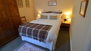 Een slaapkamer met een bed, twee nachtkastjes met lampen, een houten garderobe en een geruite deken op het bed.