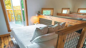 A loft-style bedroom with a bed, wooden beams, a window, and a view of the outdoors.