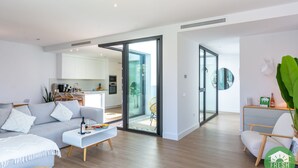 A modern living room with a sofa, coffee table, and a dining area. There is a kitchen visible through an open door.