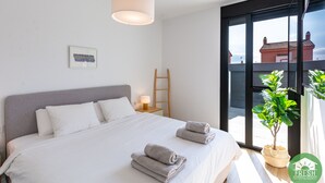 A modern bedroom with a large bed, a bedside table with a lamp, a framed picture on the wall, a ladder, and a potted plant by the window.