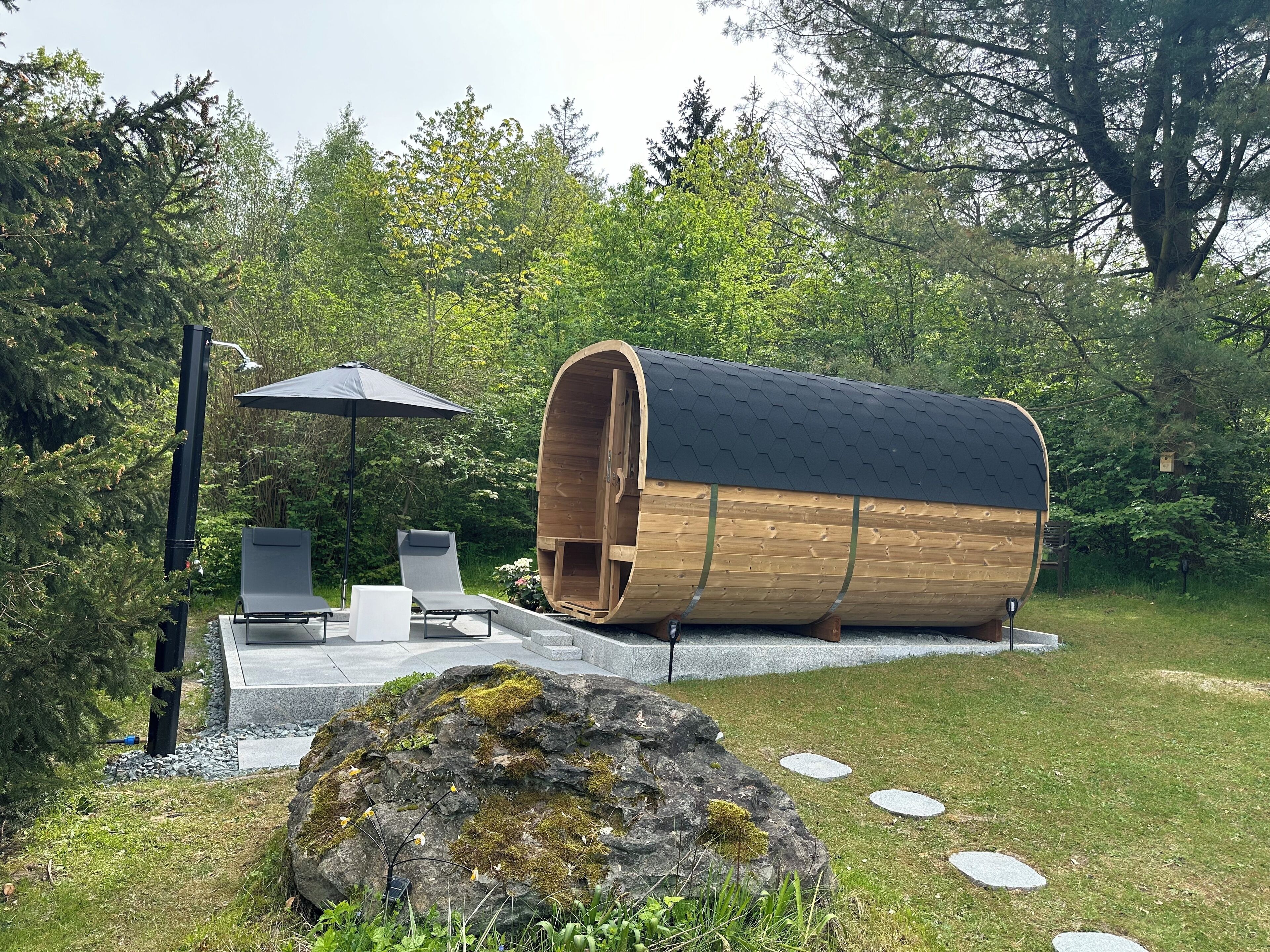 A wooden barrel-shaped structure with a door, surrounded by a garden with a rock, a bench, and an umbrella.