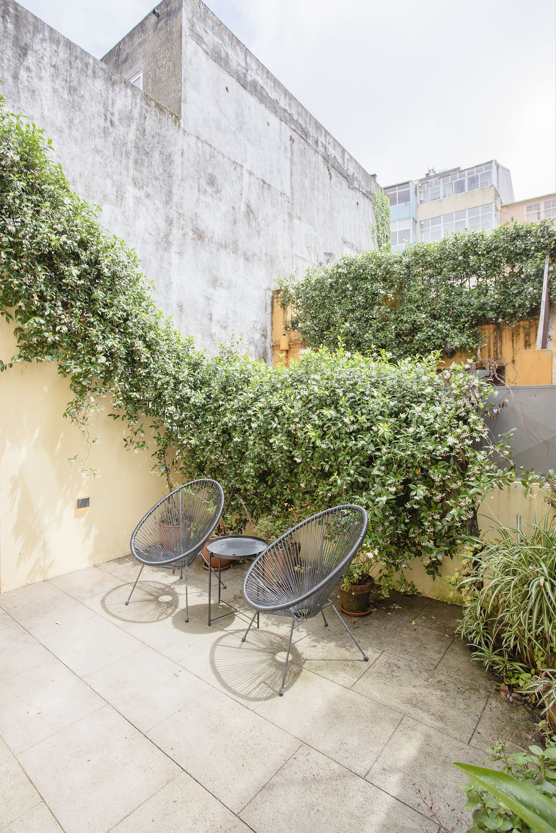 A patio with two metal chairs and a small table, surrounded by greenery and a concrete wall.