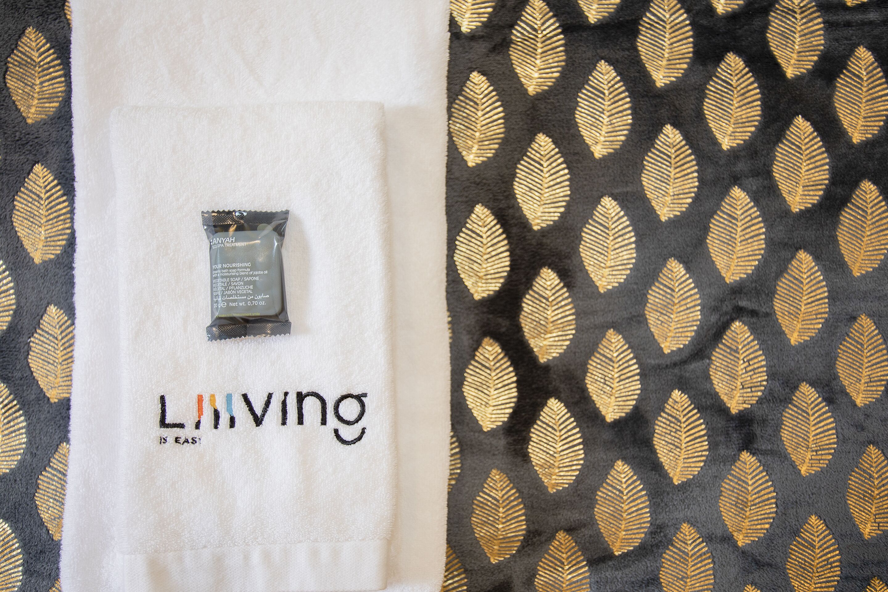 A white towel with the text "LIVING IS EASY" and a small packet labeled "ANYAH" on a black fabric with a gold leaf pattern.