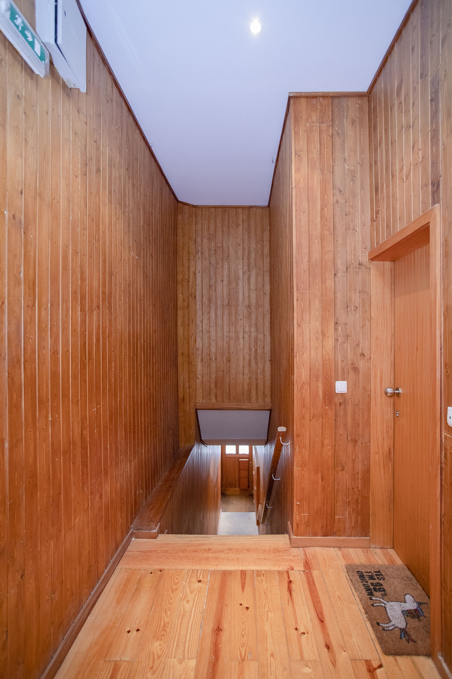 A wooden interior with a central staircase, a door on the right, and a small table at the top of the stairs.