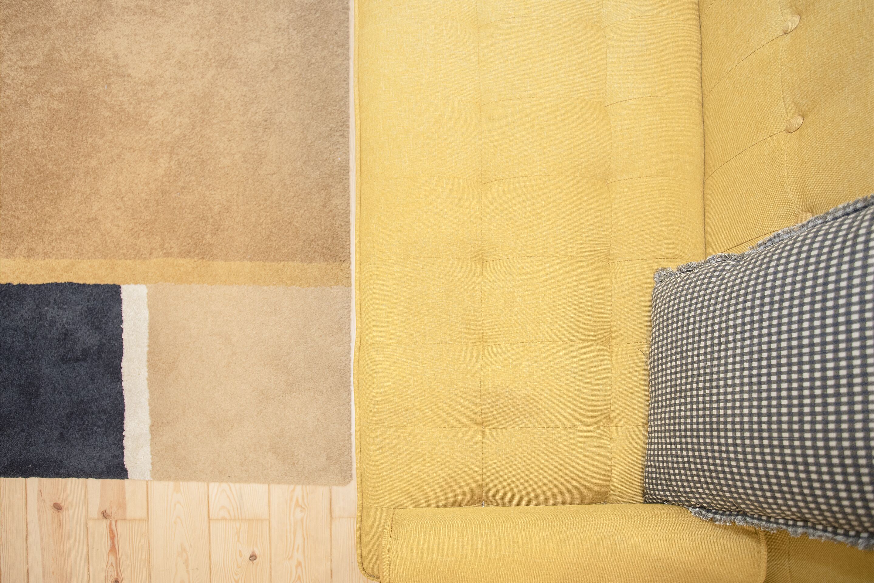 A corner of a room with a yellow upholstered sofa, a checkered pillow, and a multi-colored area rug.