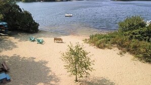 Una playa arenosa con un banco y dos sillas verdes, un árbol pequeño y un bote en el agua.