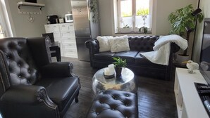 Ein Wohnzimmer mit schwarzem Ledersofa, einem passenden Sessel und einem Couchtisch. Es gibt einen weißen Kühlschrank, eine weiße Schrank und ein Fenster mit Blick auf Grünflächen.