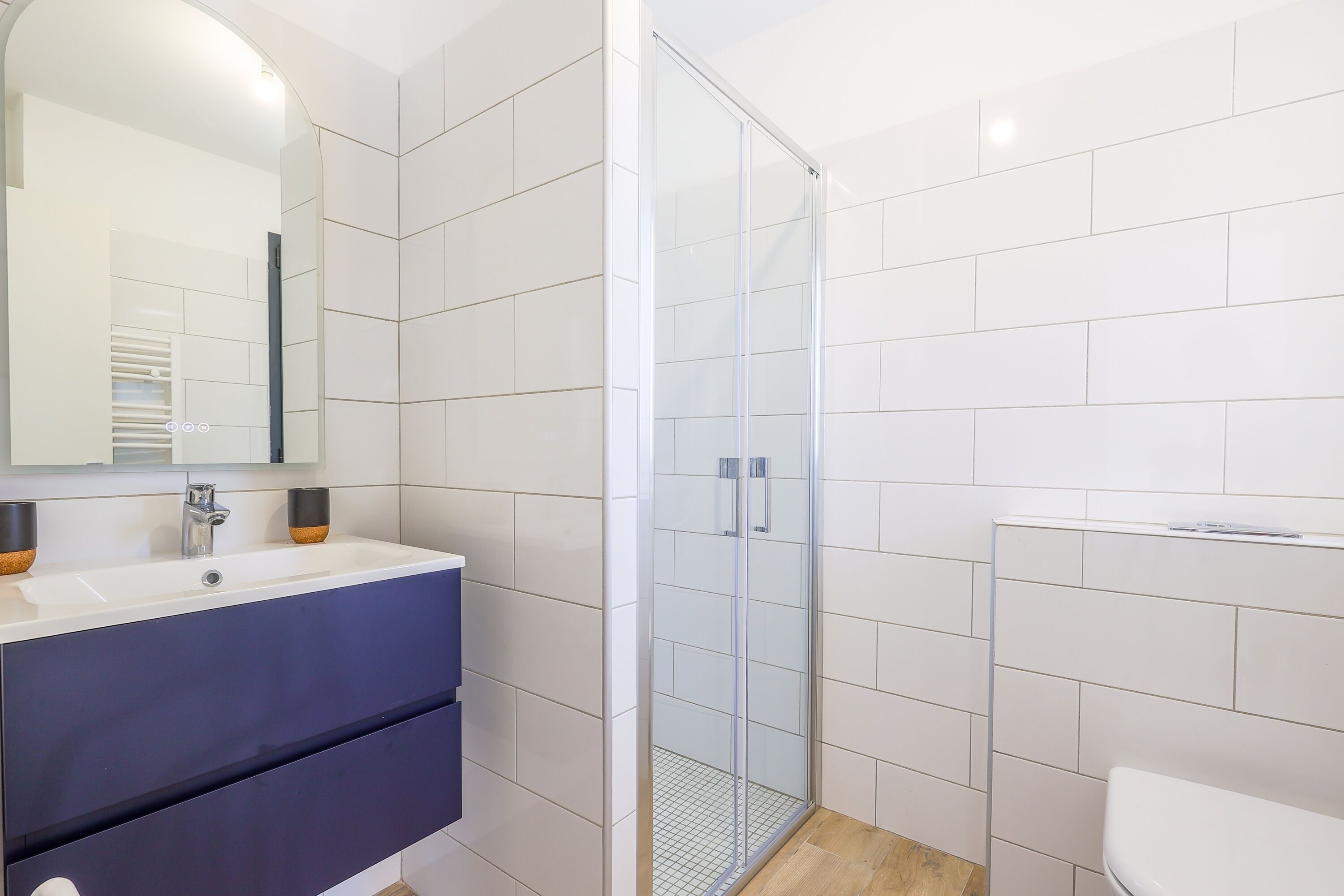 Un baño moderno con un lavamanos blanco, tocador azul oscuro, cabina de ducha con mampara de vidrio y paredes cubiertas de azulejos blancos.