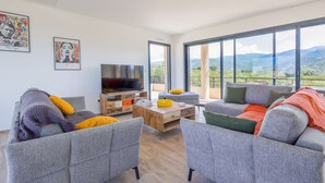A modern living room with grey sofas, a wooden coffee table, and a flat-screen TV. There are large windows offering a view of the outdoors. - Villa Lurenzu - Maison pour 8 avec piscine (Belgodère)