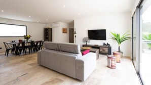 Ein modernes Wohnzimmer mit grauem Sofa, Esstisch und Stühlen. Ein Flachbildfernseher ist an der Wand befestigt, es gibt einen Blumentopf und eine Glasschiebetür, die auf einen Balkon führt.