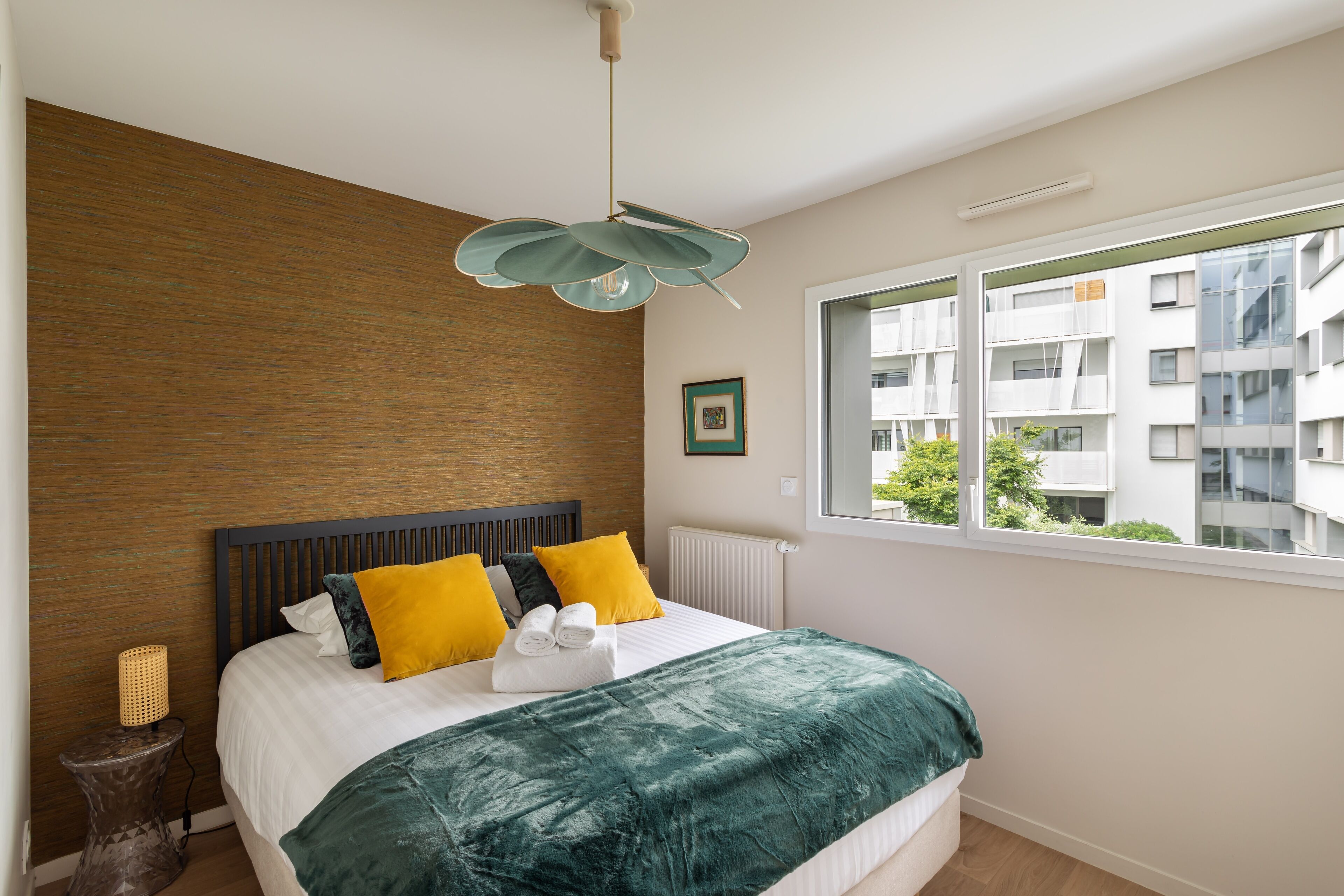 Quarto moderno com parede de destaque em madeira, uma cama grande com almofadas amarelas e verdes, e vista para a cidade através de uma janela ampla.